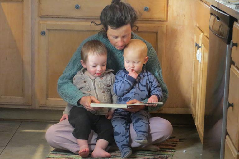 Woman holding two infants on the kitchen floor reading a book to them